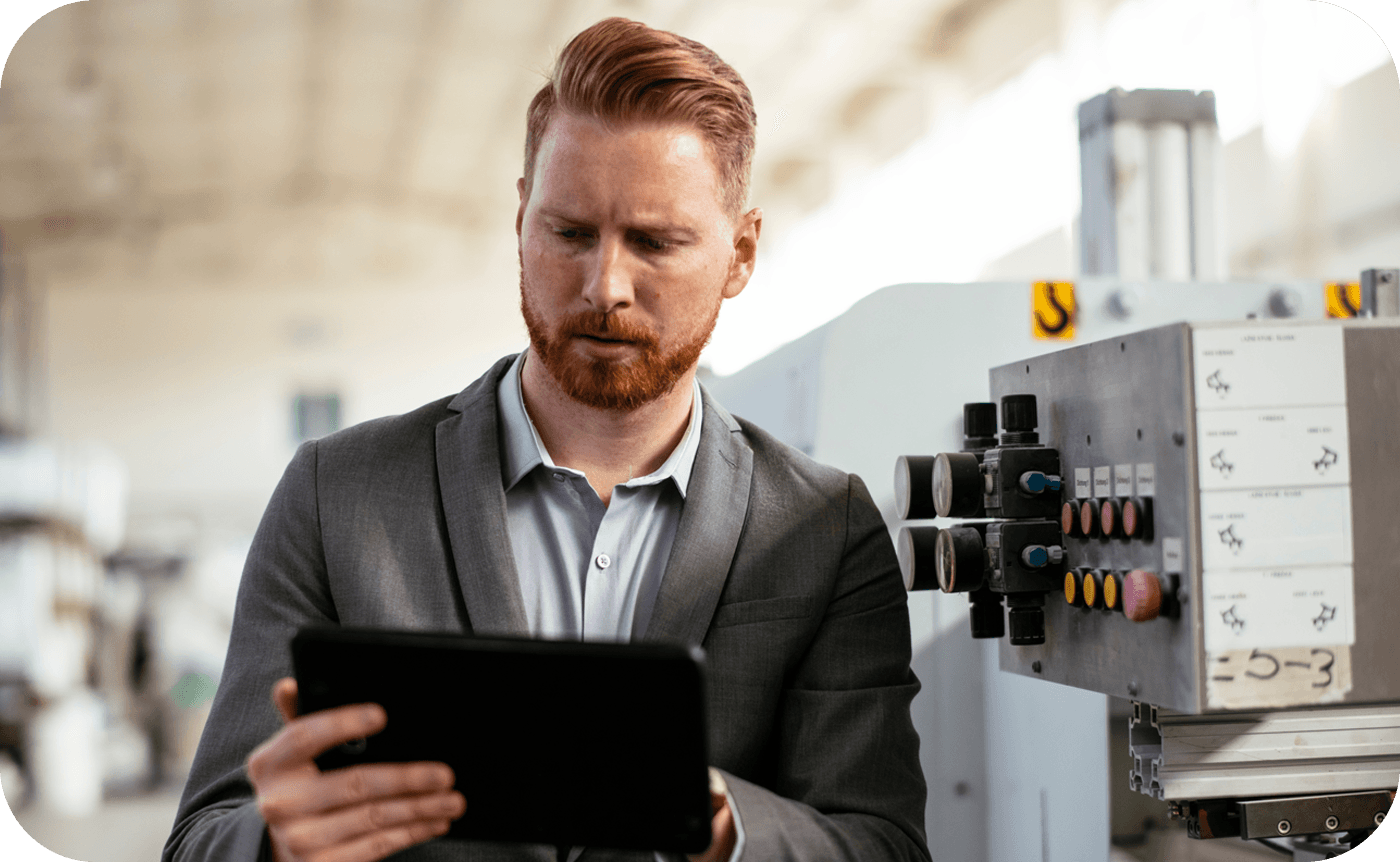 A man in a suit examines a tablet while standing next to industrial machinery in a spacious, brightly lit factory setting.