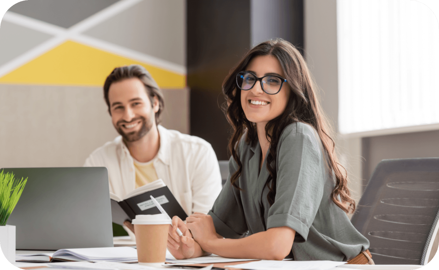 Two people sitting at a table, one holding a book, the other smiling towards the camera, in a modern office with abstract wall art and a laptop nearby.