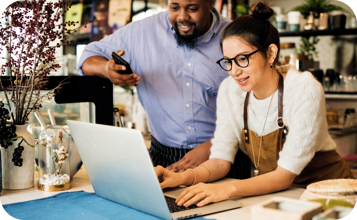 A woman wearing glasses and an apron types on a laptop, while a man in a blue shirt watches, in a cozy café surrounded by plants and coffee equipment.