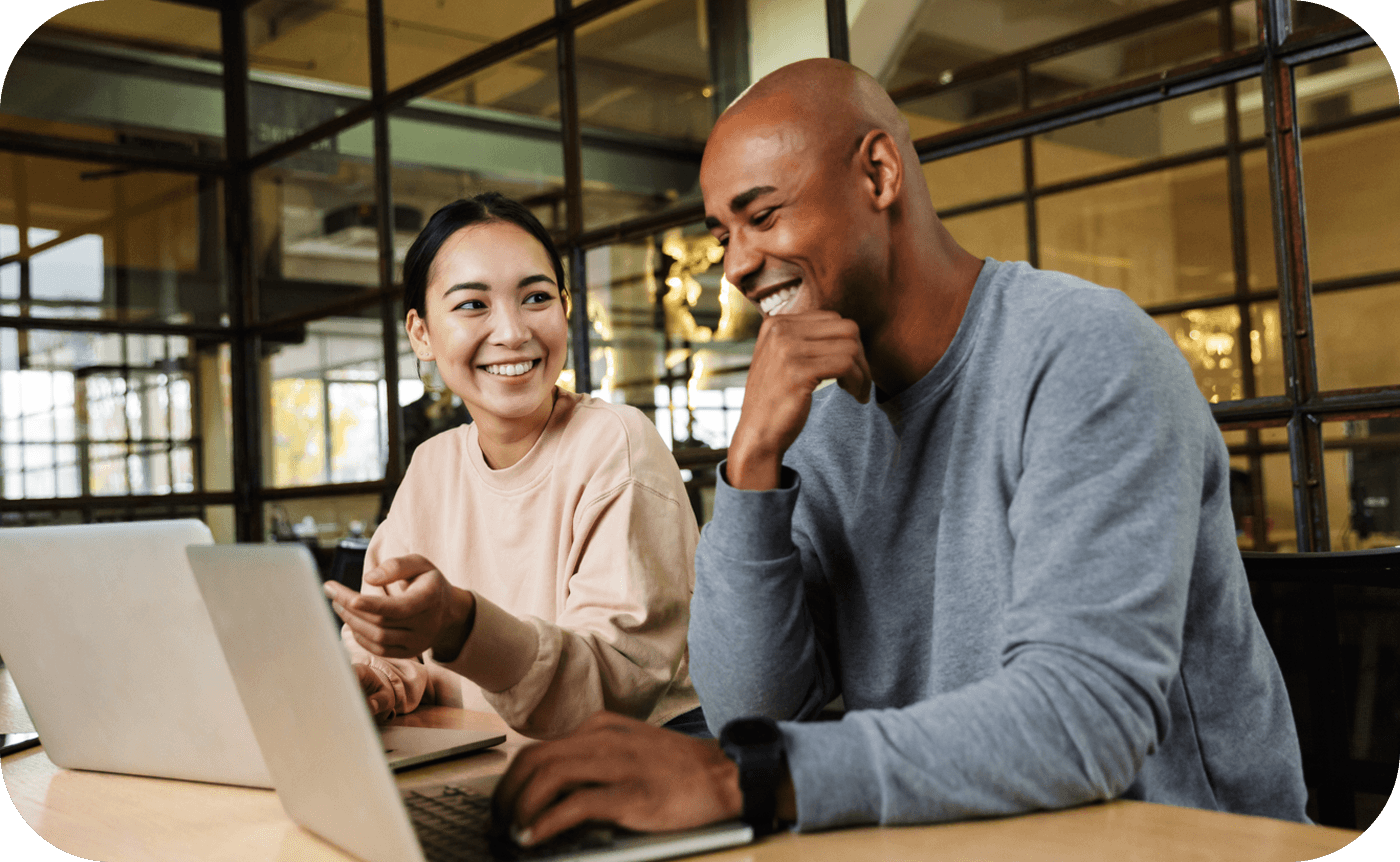 Two people smile while working on laptops in a modern office with glass walls and ambient lighting.