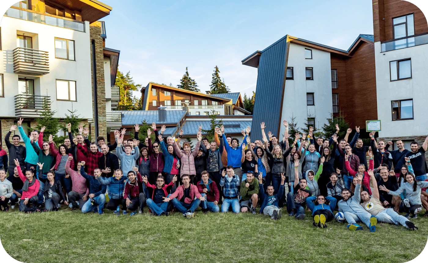 A large group of people sit and stand together, raising their hands in celebration, surrounded by modern buildings and green grass, under a clear blue sky.