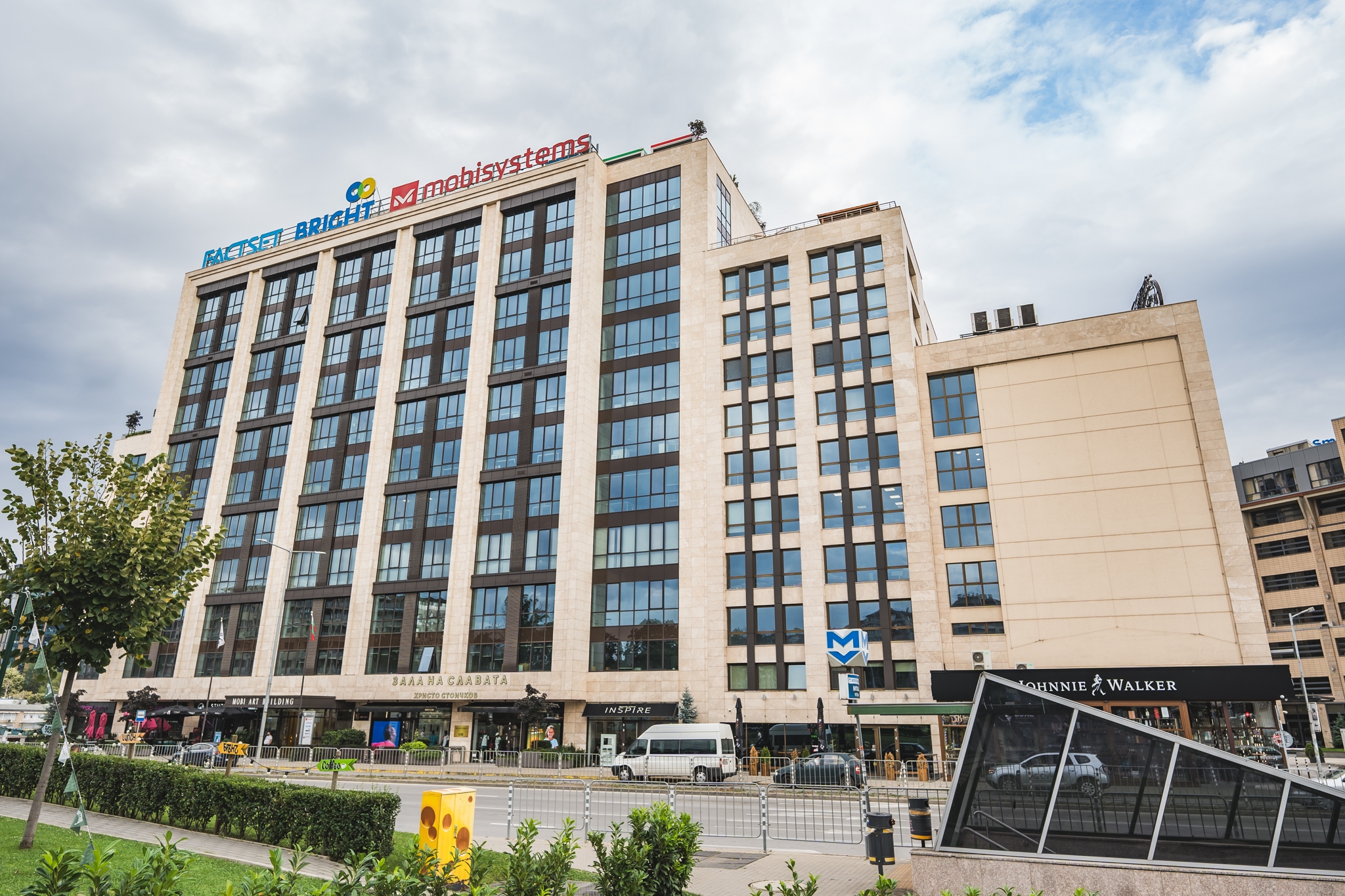 A tall building with "MobiSystems," "Racket," and "Brighte" signs stands beside a road. Vehicles and pedestrians move below, while greenery and a metro entrance enhance the urban setting.