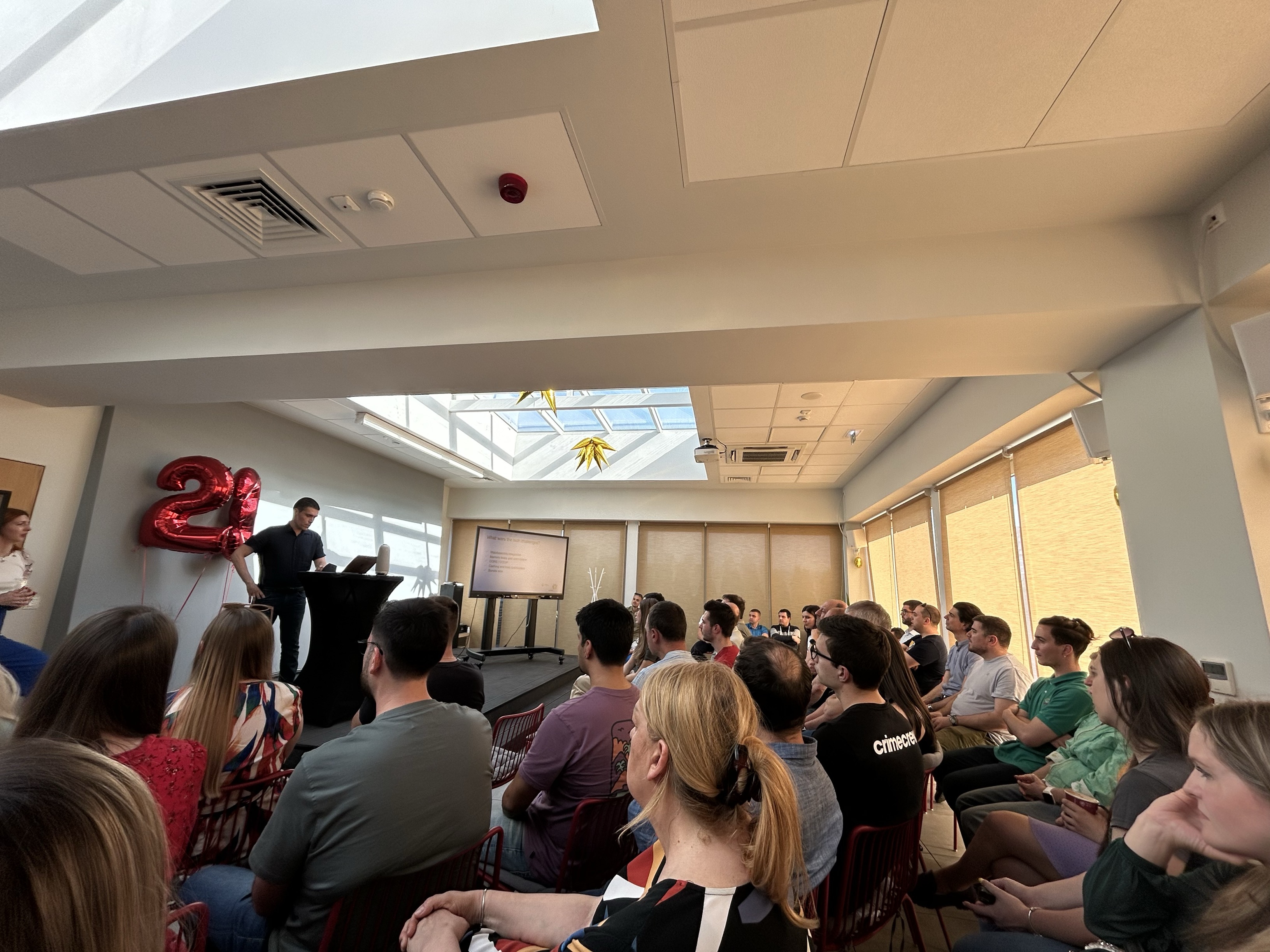 A speaker presents to an attentive audience in a well-lit room, with "21" balloons and a screen displaying a presentation in the background.