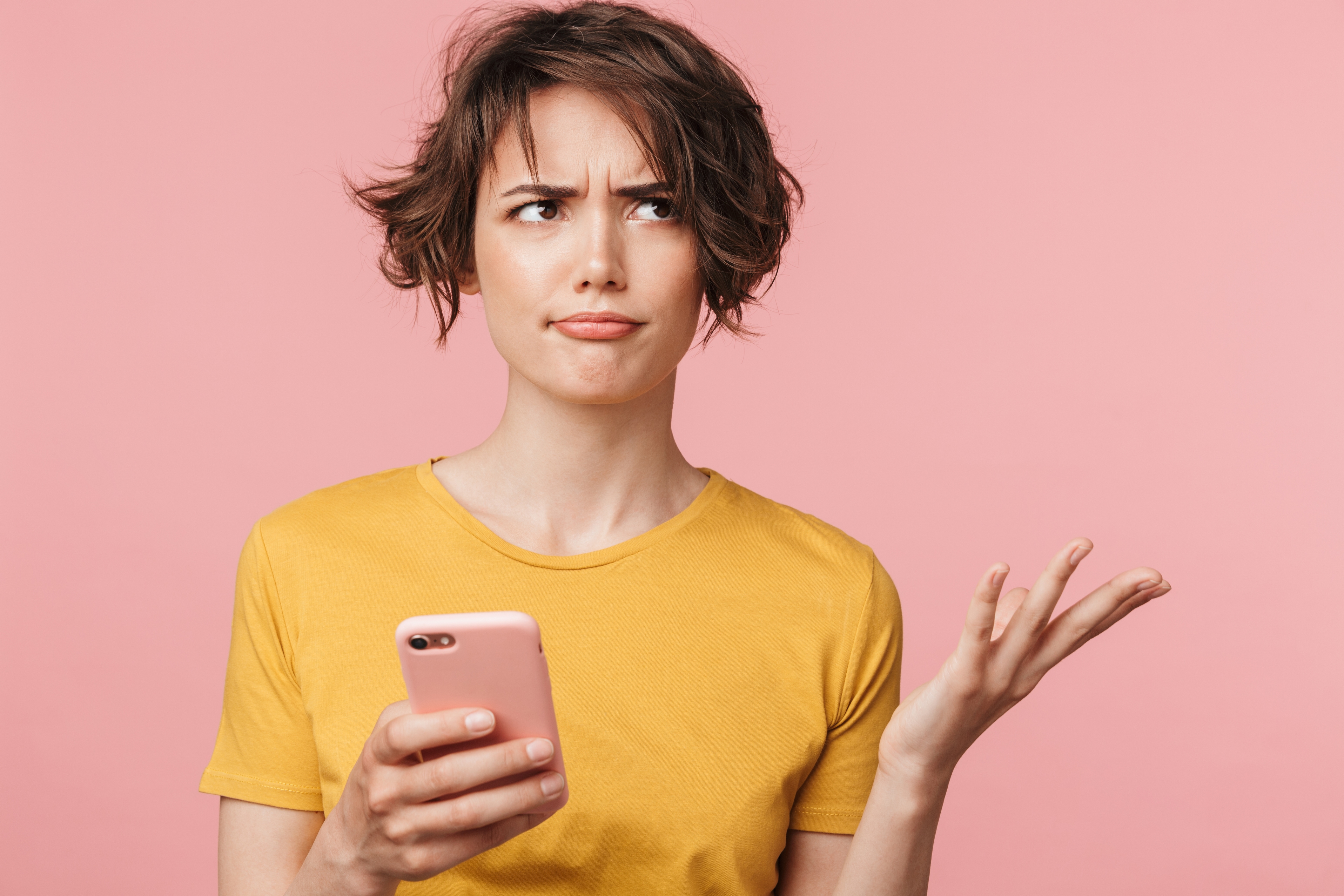 Woman with short brown hair in yellow t-shirt looking confused and frustrated while holding pink smartphone against pink background.